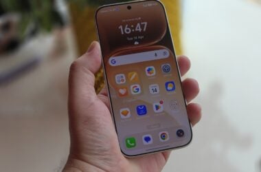 Hand holding an Android phone showing its home screen in front of window blinds